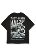 An Oversized t-shirt featuring a large white and gray print on the back. The graphic includes the text 'THE THINKER' and its definition above a grayscale sculpture image with wavy black lines, and smaller white text below. Design by bakaka.